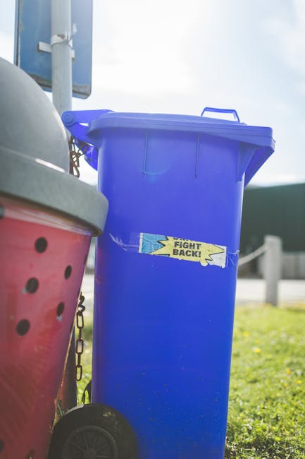 A large collection of mixed waste, including crumpled cardboard boxes, plastic bags, paper, and assorted packaging materials, is piled on a paved area beside a street. The waste is overflowing from a