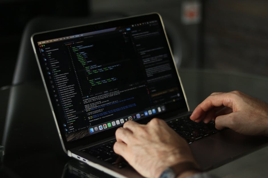 A person seated at a desk in an indoor setting, working on a laptop displaying a screen filled with multicolored lines of code, likely related to programming or software development. The individual is